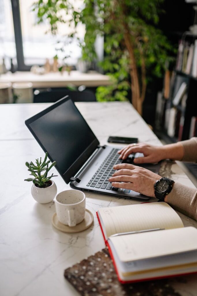 pexels photo 6818307 Adult typing on a laptop surrounded by indoor plants, notebooks, and a cup in a modern workspace.